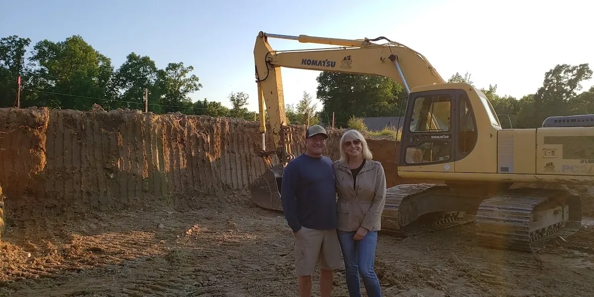 Two people stand smiling in front of a yellow Komatsu excavator at a construction site with dirt and trees in the background.
